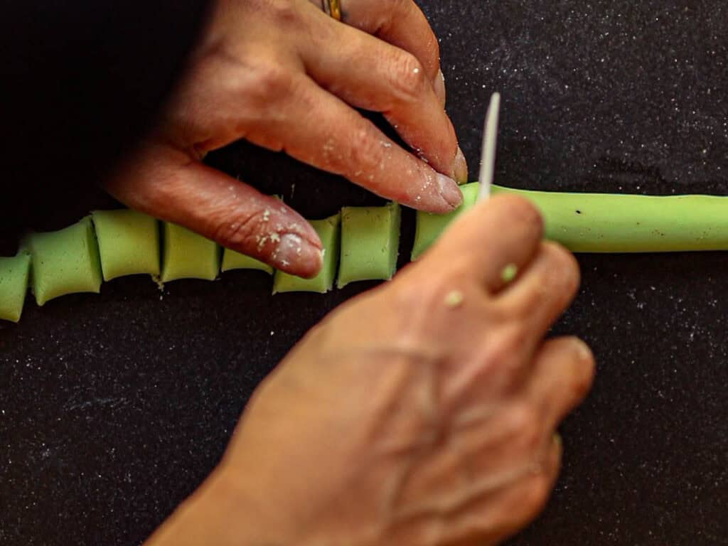 Close-up of hands shaping and cutting green dough on a dark surface. One hand is pinching and the other is holding a tool, making even, small sections in the clay. The focus is on the detailed work and texture.